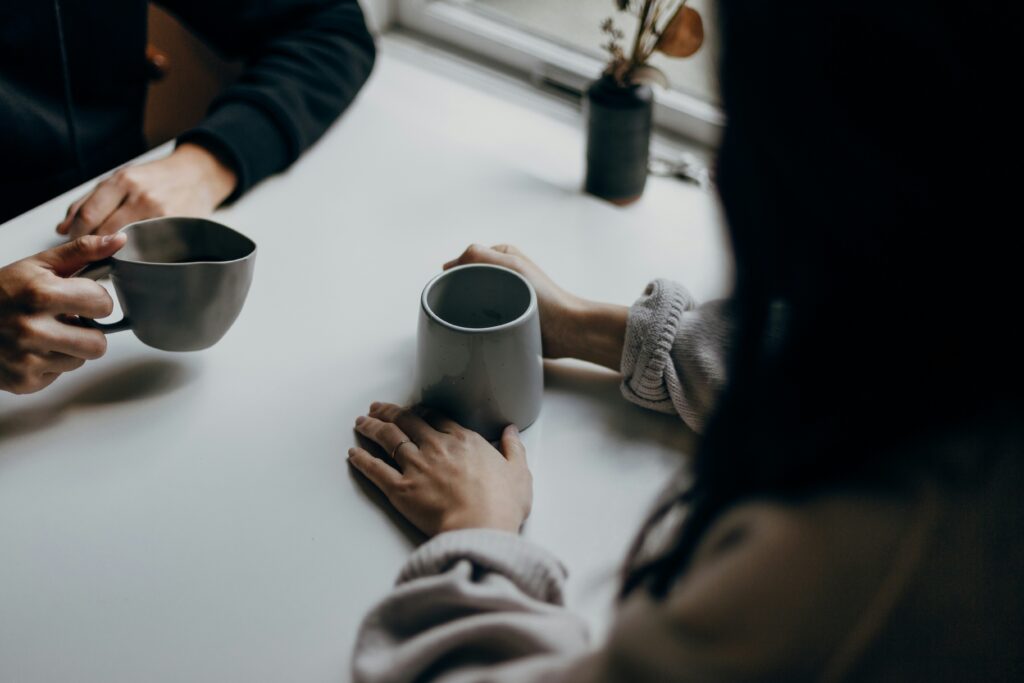 Manos de dos personas con tazas de café sobre una mesa junto a una ventana, en medio de una conversación.