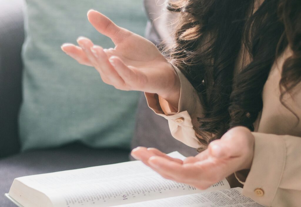 Manos de una mujer en el aire, con las palmas hacia arriba, como explicando algo, mientras un libro abierto descansa sobre sus piernas.