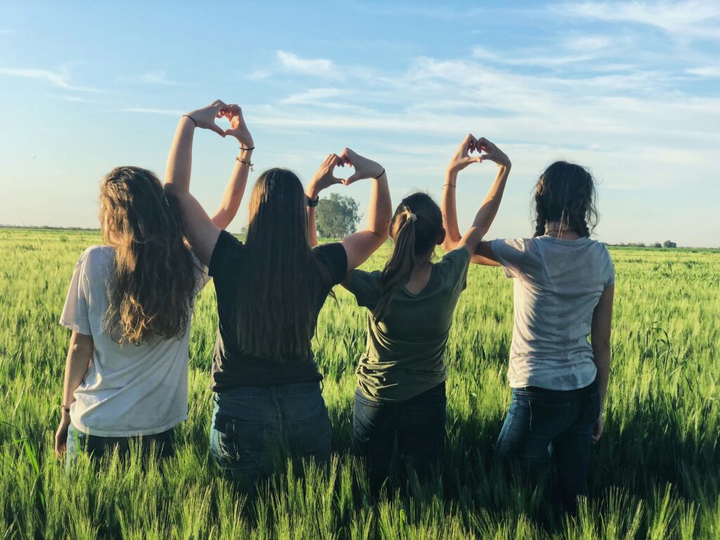 Cuatro mujeres de espaldas, con los brazos entrelazados y formando corazones con las manos, en un campo verde bajo un cielo azul.